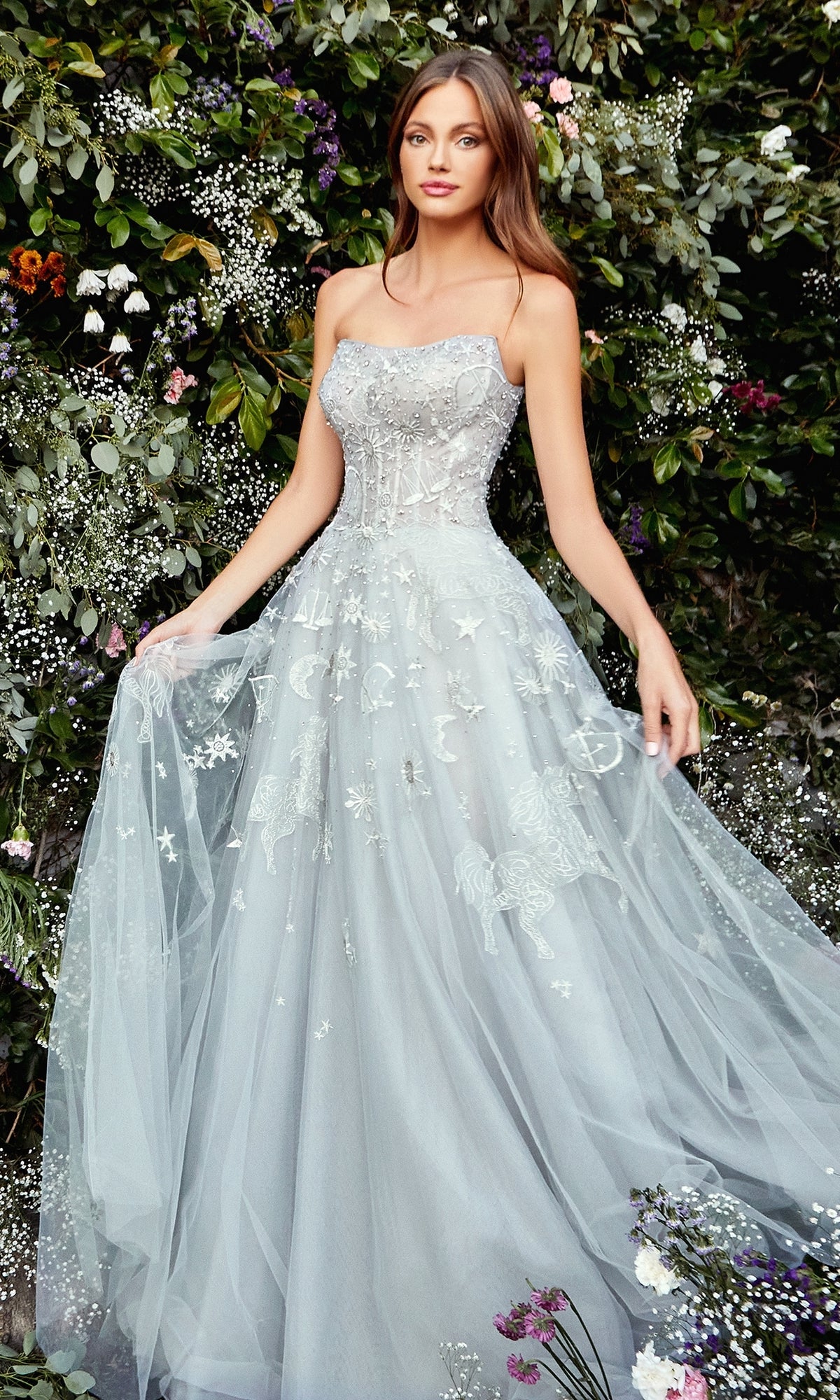 A woman wearing the Constellation-Print Strapless Prom Ball Gown A0890 poses before a wall adorned with green foliage and small flowers.